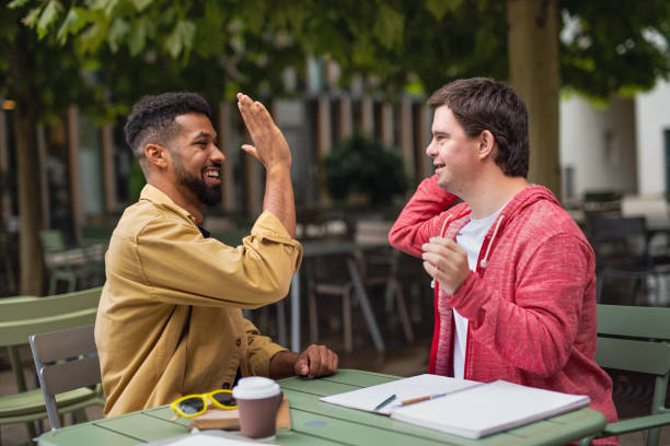 istockphoto-1349910331-612x612 Young man with Down syndrome with his mentoring friend sitting outdoors in cafe high fiving.